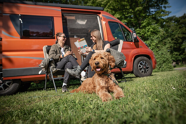 Camping Neuhof a.d.Zenn Zwei Personen und ein Hund sitzend vor einem organgefarbenen Camper