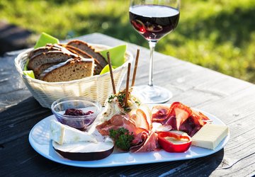 Florian Trykowski Teller mit Wurst, Käse, Brot und Marmelade sowie ein Glas Rotwein auf einem Holztisch im Grünen – angerichtet für eine Brotzeit im Freien.
